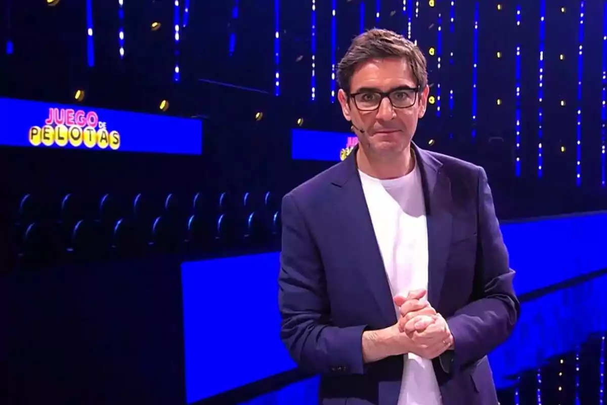 Juanra Bonet con gafas y chaqueta azul está de pie en un set de televisión iluminado con luces azules y el letrero de Juego de Pelotas al fondo Juanra Bonet con gafas y chaqueta azul está de pie en un set de televisión iluminado con luces azules y el letrero de Juego de Pelotas al fondo