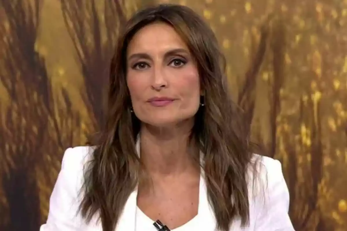 Leticia Iglesias de cabello castaño largo y suelto con chaqueta blanca posando frente a un fondo marrón difuminado en Informativos Telecinco