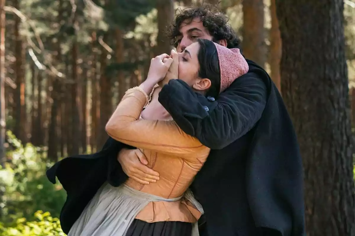 Un home abraça fortament Loren Mairena tapant-li la boca amb la mà en un bosc, tots dos porten roba d’època a Valle Salvaje. Un home abraça fortament Loren Mairena tapant-li la boca amb la mà en un bosc, tots dos porten roba d’època a Valle Salvaje.