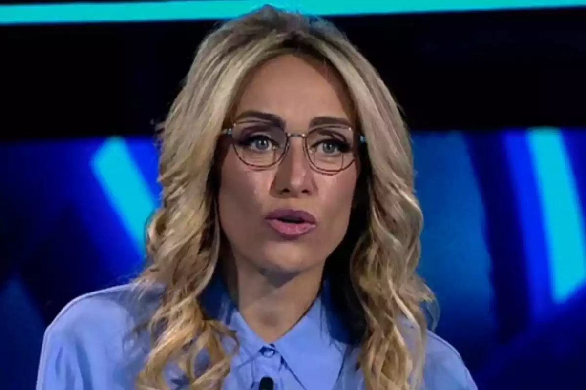 Luján Argüelles de cabello rubio y ondulado con gafas y camisa azul hablando en un estudio de El rival más débil con luces azules de fondo.