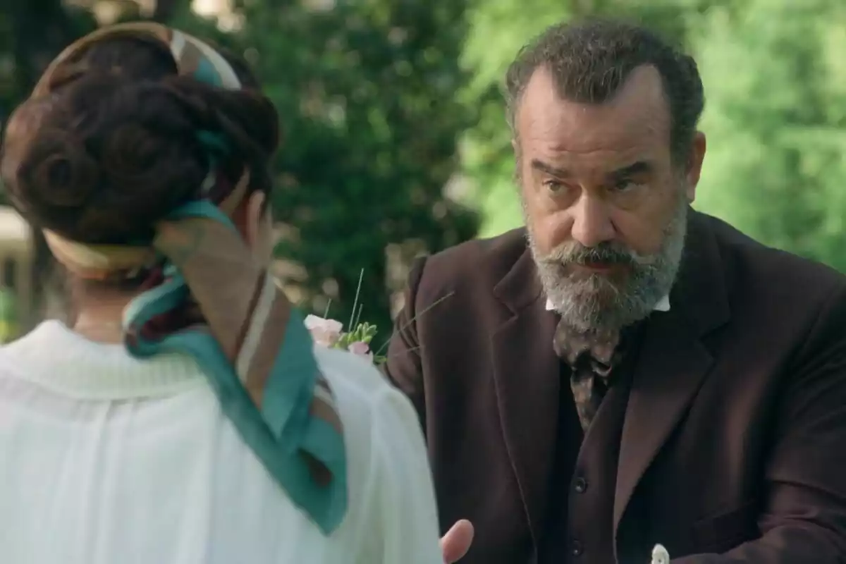Manuel Regueiro de barba canosa y traje oscuro conversa con Carmen Asecas de espaldas que lleva un pañuelo en el cabello en un entorno al aire libre con vegetación de fondo en La Promesa Manuel Regueiro de barba canosa y traje oscuro conversa con Carmen Asecas de espaldas que lleva un pañuelo en el cabello en un entorno al aire libre con vegetación de fondo en La Promesa