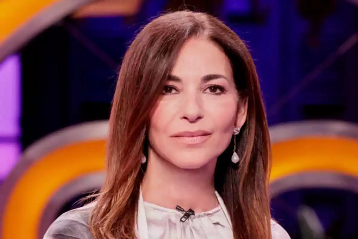 Mariló Montero de cabell castany llis i llarg, amb arracades de perla, vestida amb una brusa clara i fons desenfocat en tons morats i grocs a MasterChef Celebrity