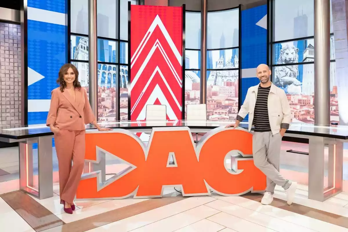 Marta Flich i Gonzalo Miró posen somrients en un modern plató de Directe al gra amb un gran rètol taronja que diu DAG i pantalles de fons amb gràfics i paisatges urbans. Marta Flich i Gonzalo Miró posen somrients en un modern plató de Directe al gra amb un gran rètol taronja que diu DAG i pantalles de fons amb gràfics i paisatges urbans.