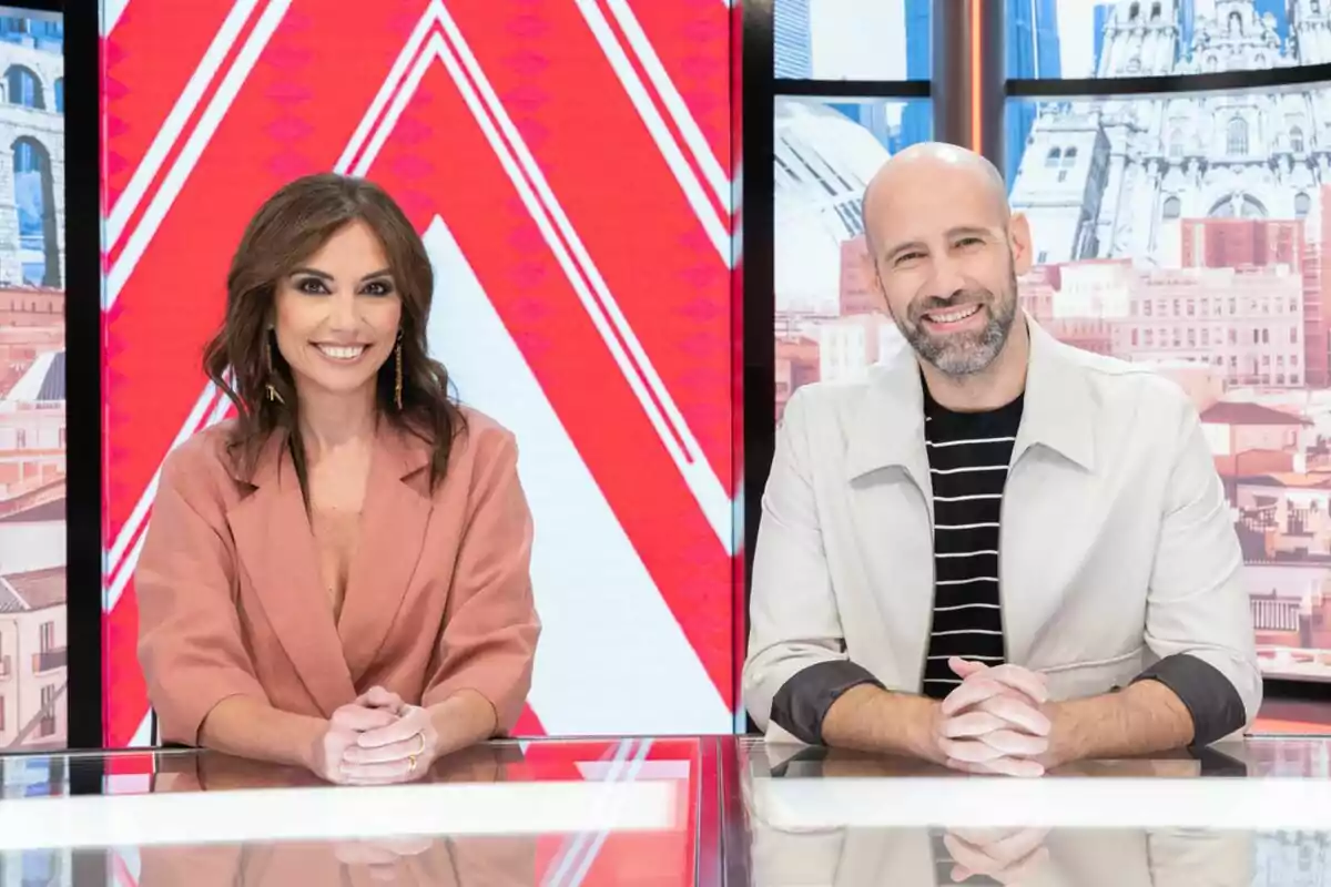 Marta Flich i Gonzalo Miró somriuen asseguts darrere d’una taula en un plató de televisió de Directe al Gra amb un fons colorit i urbà.