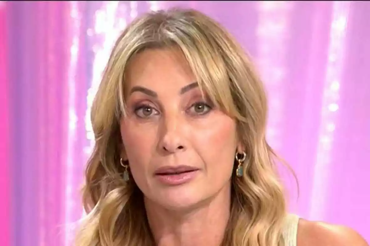 Mónica Pont rubia de cabello ondulado y largo con expresión seria frente a un fondo rosa difuminado en Fiesta