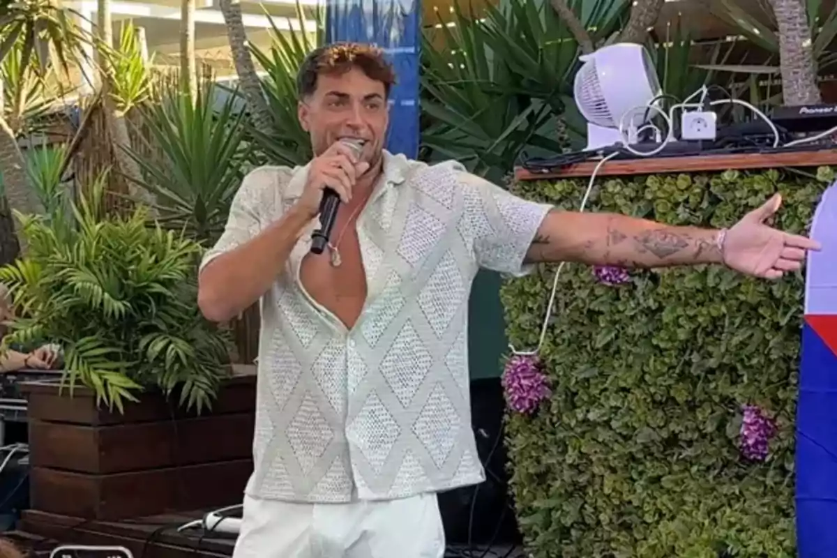 Montoya somrient amb micròfon a la mà, camisa clara oberta i fons de plantes verdes Montoya somrient amb micròfon a la mà, camisa clara oberta i fons de plantes verdes