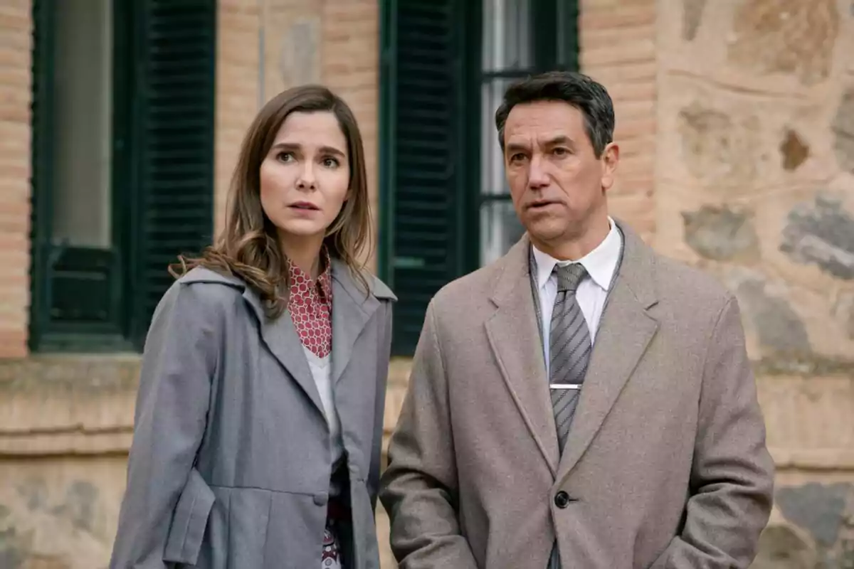 Dos personas con expresión seria y abrigos de invierno, que son Oriol Tarrasón y Natalia Sánchez, están de pie frente a un edificio de ladrillo y piedra con ventanas oscuras. Dos personas con expresión seria y abrigos de invierno, que son Oriol Tarrasón y Natalia Sánchez, están de pie frente a un edificio de ladrillo y piedra con ventanas oscuras.