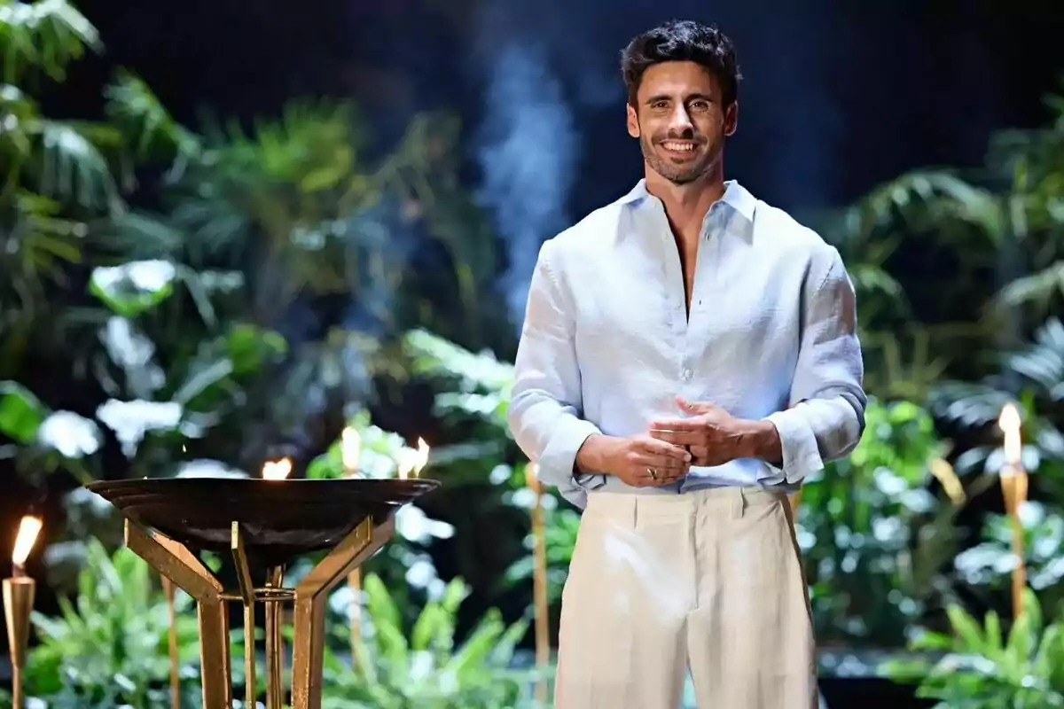 Home somrient dret amb camisa blanca i pantalons clars en un entorn tropical amb torxes i vegetació de fons Home somrient dret amb camisa blanca i pantalons clars en un entorn tropical amb torxes i vegetació de fons