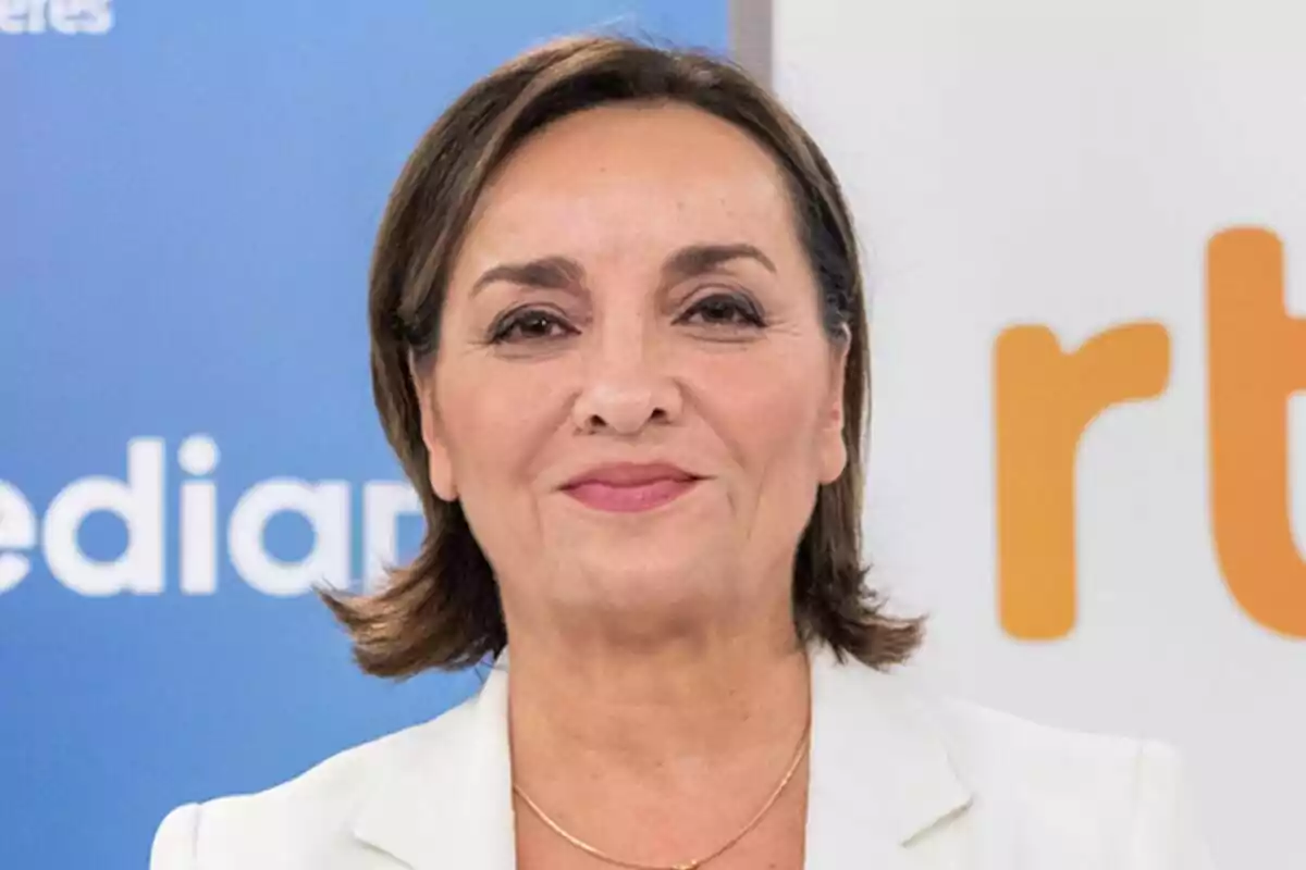Pepa Bueno de mediana edad con cabello corto y castaño, vestida con chaqueta blanca y fondo con logotipos azules y naranjas del Telediario.