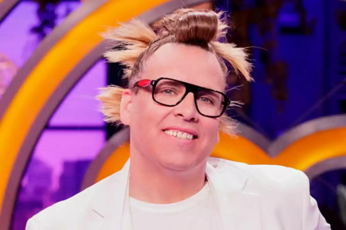 Quique Torito amb ulleres negres i pentinat cridaner amb metxes rosses cap als costats i monyo a la part superior, vestint jaqueta blanca i somrient en un ambient colorit de MasterChef Celebrity.