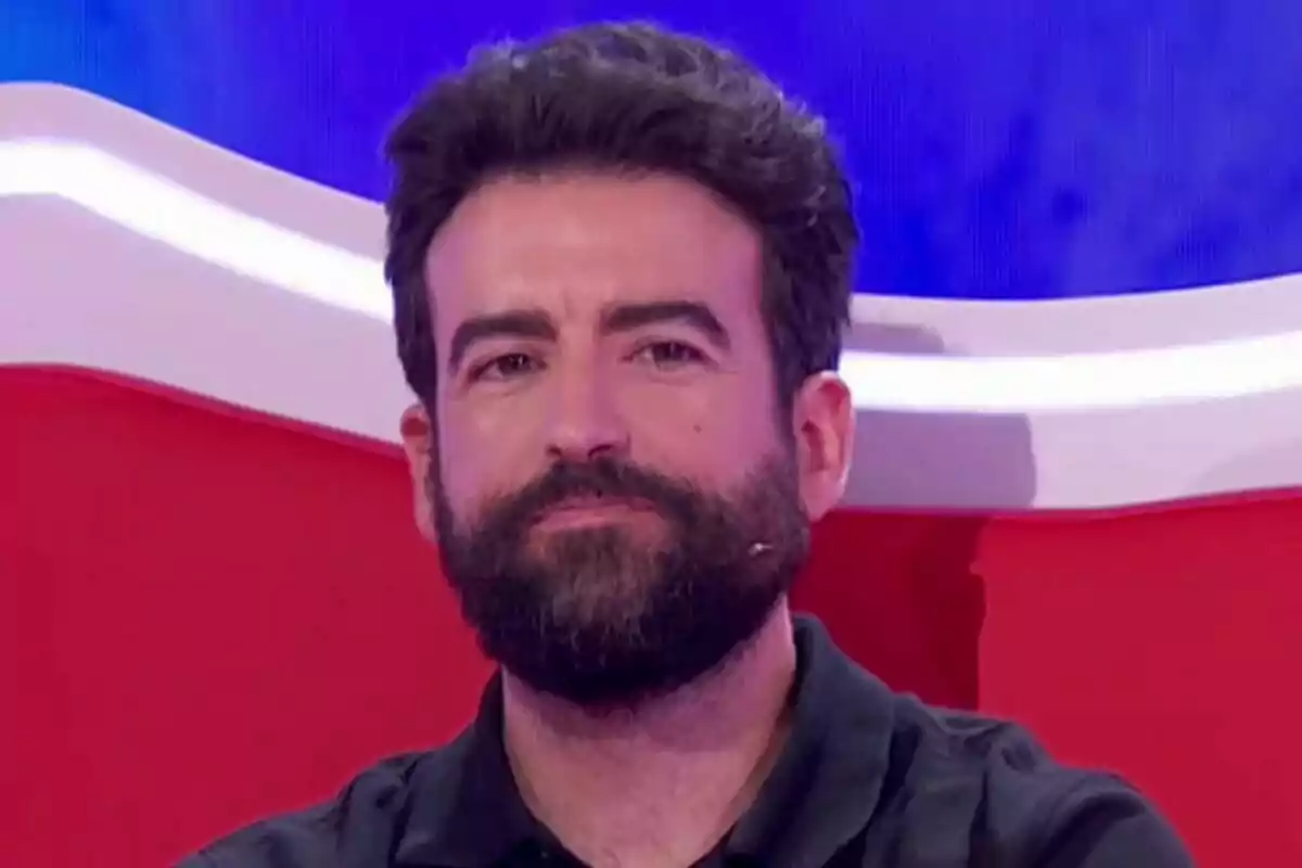 Rafa Castaño de barba y cabello oscuro sentado en un sillón rojo con fondo azul en Agárrate al sillón