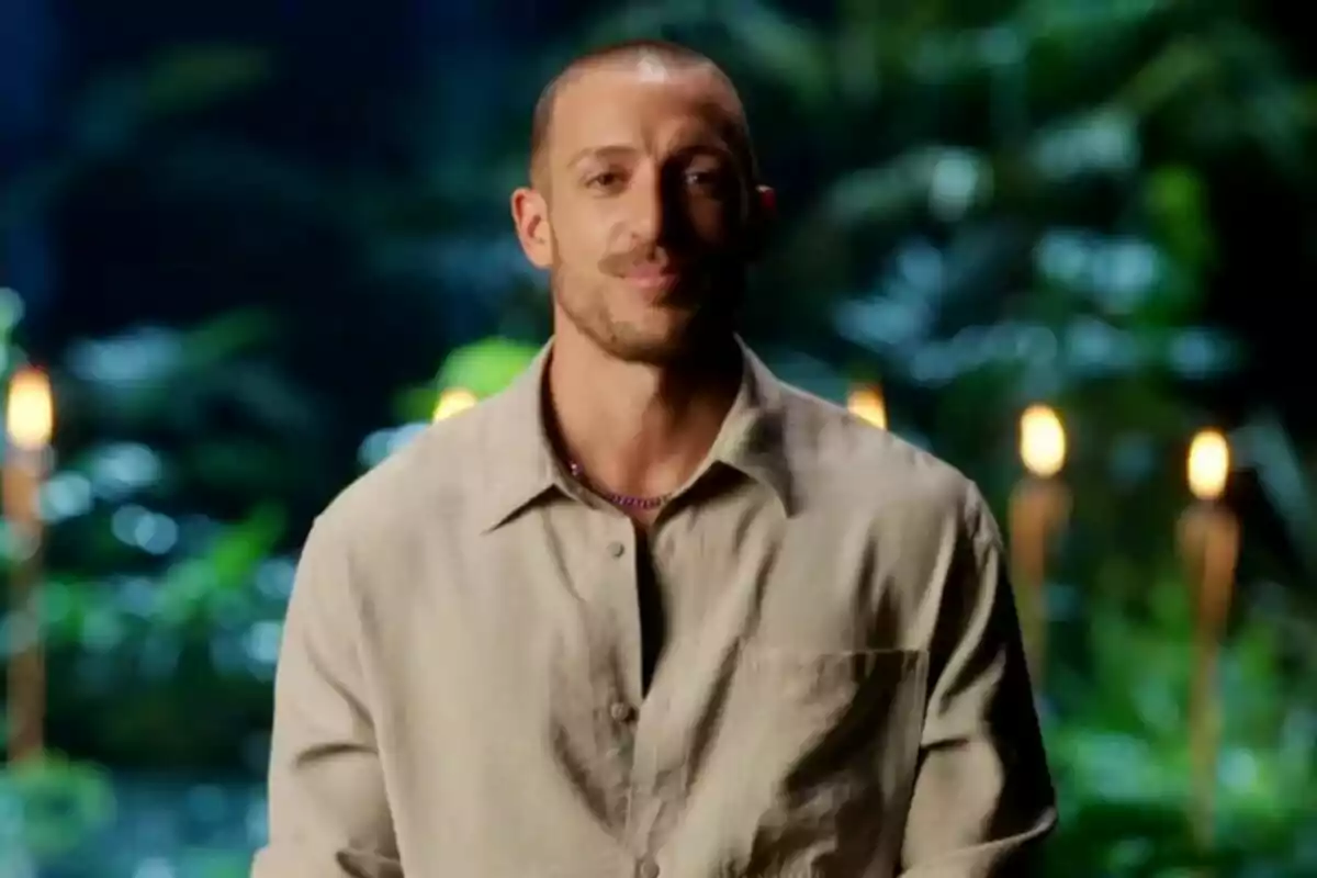 Rubén Torres, amb cabell curt i barba lleugera, porta una camisa beix i està dret davant d’un fons de vegetació amb torxes enceses a Supervivientes All Stars.