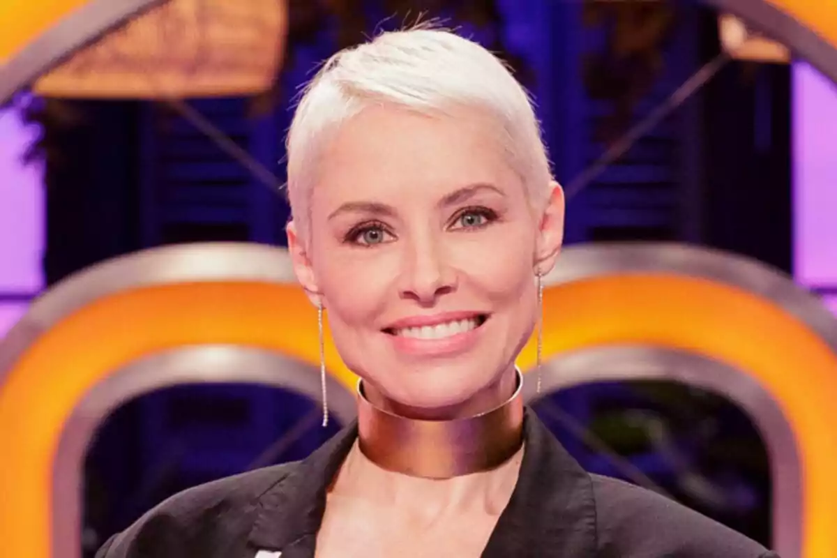 Soraya Arnelas de cabello corto y rubio sonriendo, lleva un collar ancho dorado y un fondo colorido con luces naranjas y moradas de MasterChef Celebrity