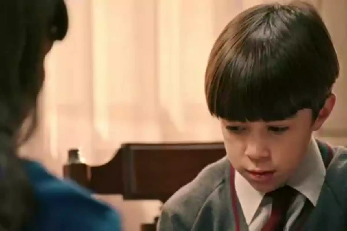Nicolás Rodicio con uniforme escolar mirando hacia abajo con expresión seria mientras está sentado en una mesa en Sueños de Libertad. Nicolás Rodicio con uniforme escolar mirando hacia abajo con expresión seria mientras está sentado en una mesa en Sueños de Libertad.