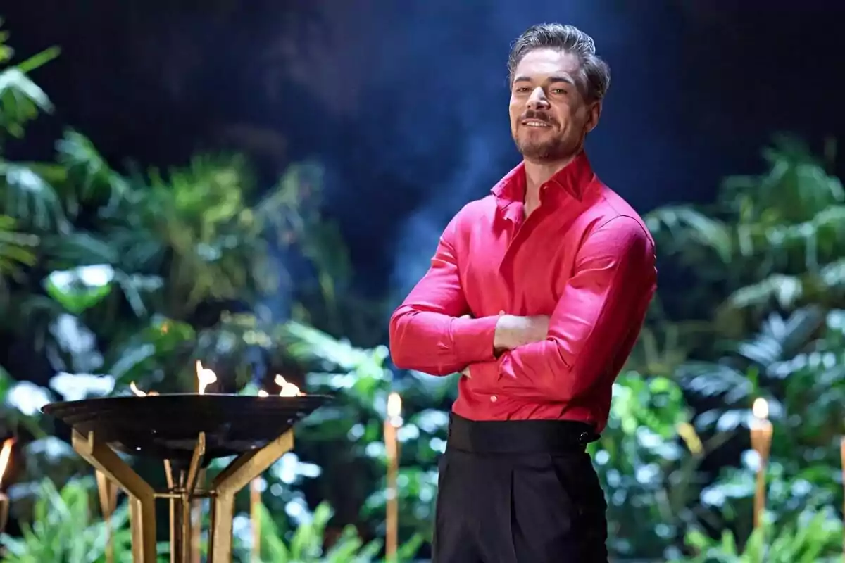 Tony Spina dret amb camisa vermella i els braços creuats en un entorn tropical amb torxes i vegetació de fons de Supervivientes. Tony Spina dret amb camisa vermella i els braços creuats en un entorn tropical amb torxes i vegetació de fons de Supervivientes.