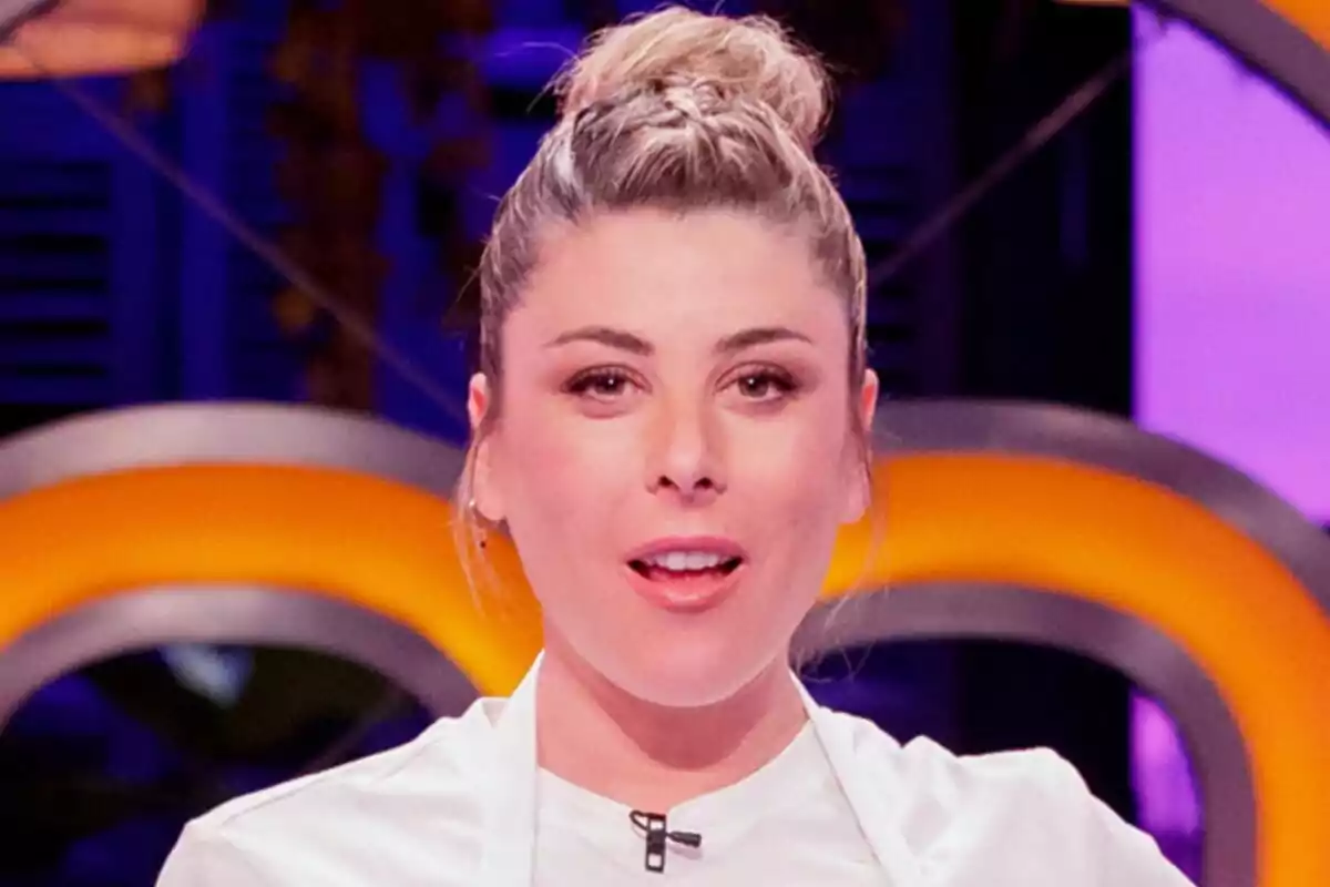 Valeria Ros joven con el cabello recogido en un moño y vestida con chaqueta blanca de chef en un set de MasterChef Celebrity con luces moradas y amarillas de fondo