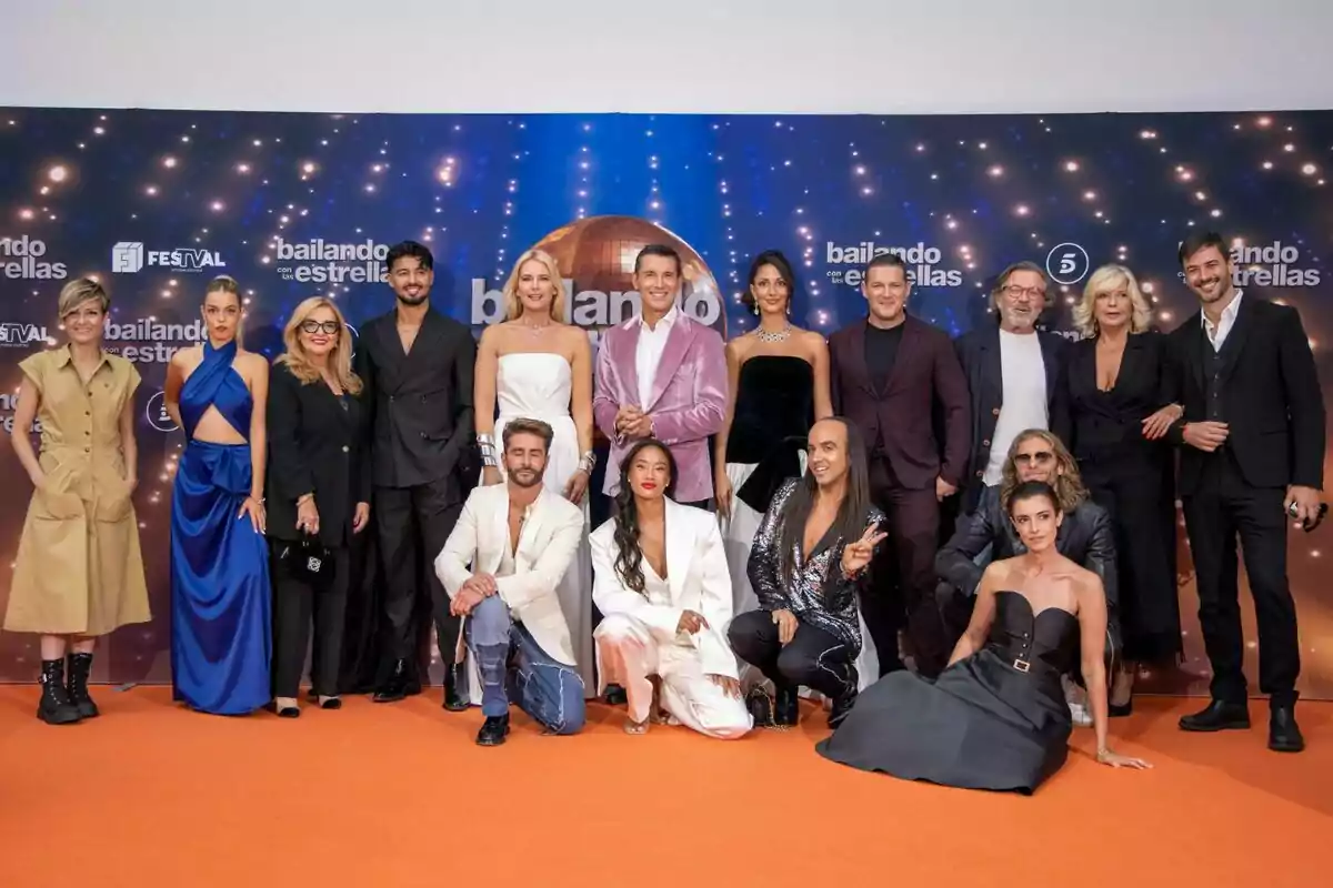 Un grupo de personas elegantemente vestidas posando sobre una alfombra naranja frente a un fondo decorado con luces y el logo de un programa de televisión llamado bailando con las estrellas