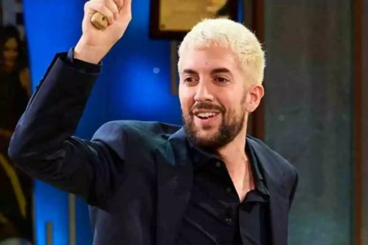 David Broncano de cabello rubio y barba oscura sonríe mientras levanta el brazo sosteniendo un objeto pequeño en un ambiente interior iluminado en La Revuelta.