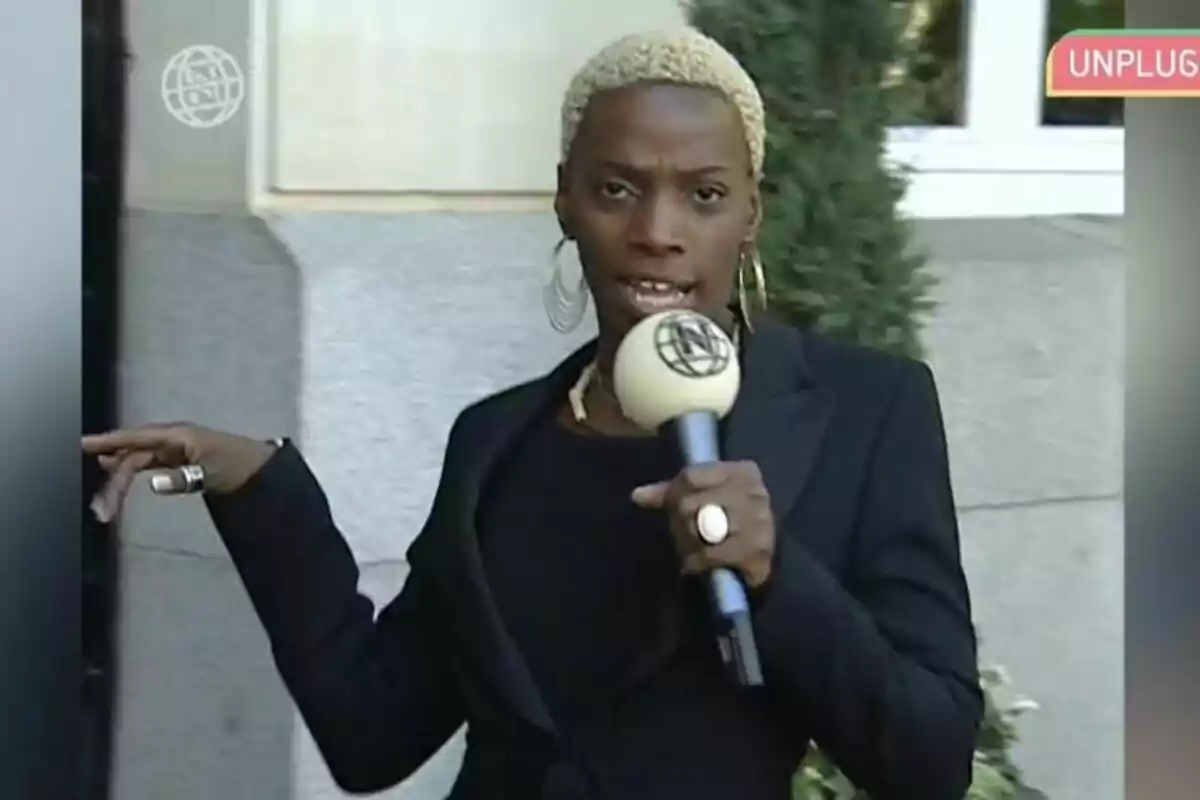 Dnoé Lamiss de cabello corto y rubio sosteniendo un micrófono mientras habla frente a una cámara, vestida con ropa oscura y accesorios llamativos, en un entorno urbano. Dnoé Lamiss de cabello corto y rubio sosteniendo un micrófono mientras habla frente a una cámara, vestida con ropa oscura y accesorios llamativos, en un entorno urbano.