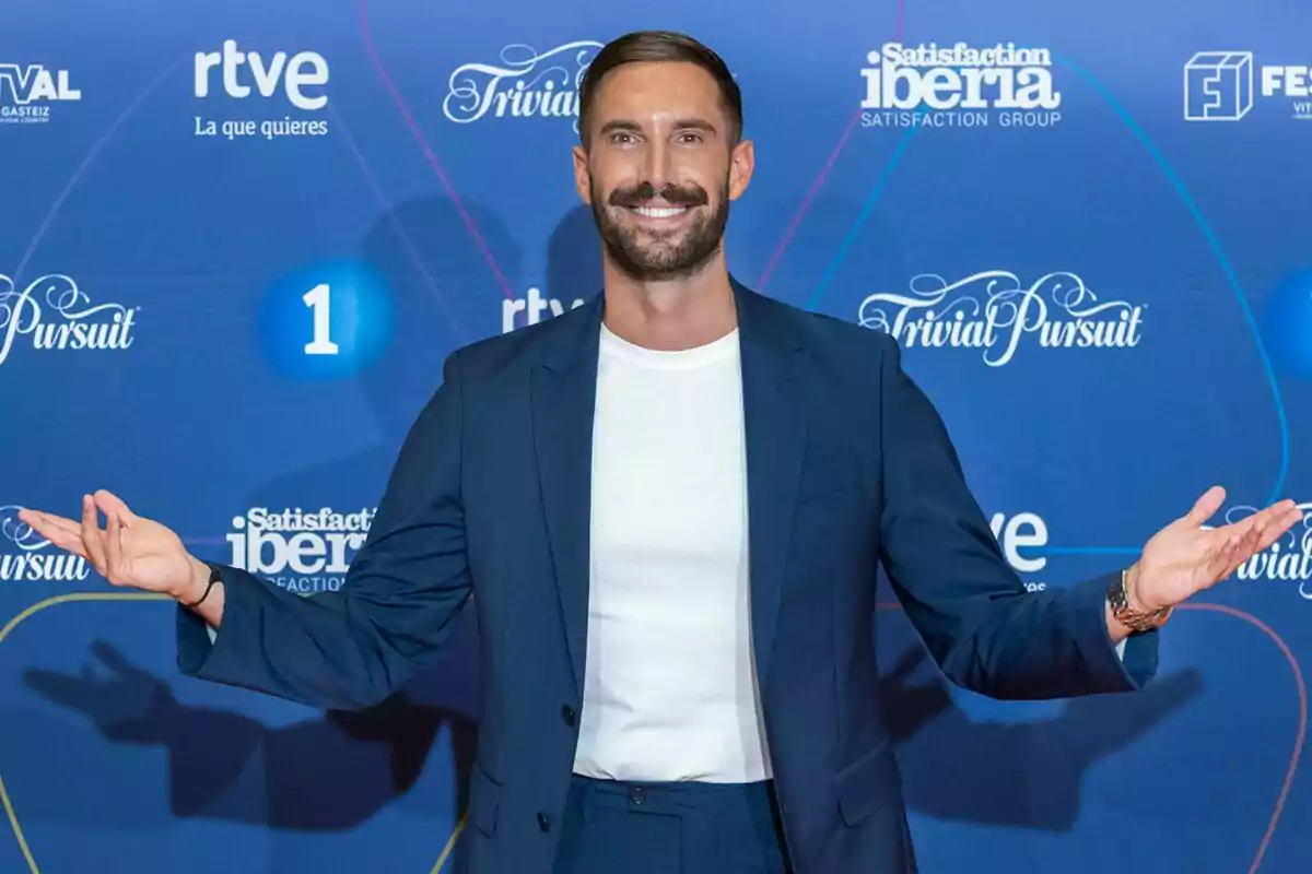 Egoitz Txurruka, ‘Txurru’, amb barba i bigoti somriu amb els braços oberts vestint un vestit blau i samarreta blanca davant d’un fons blau amb logotips de Trivial Pursuit i RTVE.