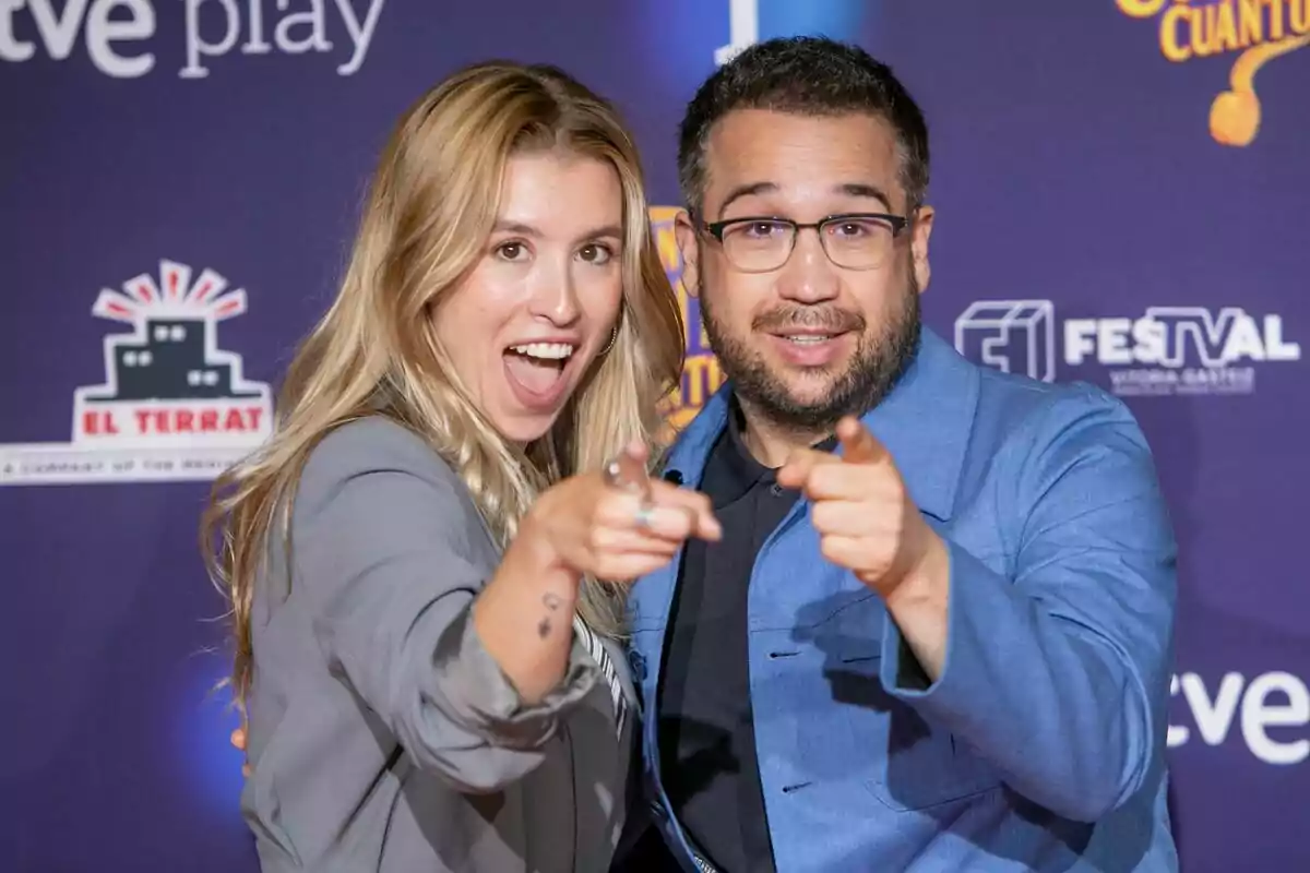 Eva Soriano y Anibal Gómez sonrientes señalando hacia la cámara en un evento con fondo azul y logotipos de patrocinadores en Cuánto cuánto cuánto.