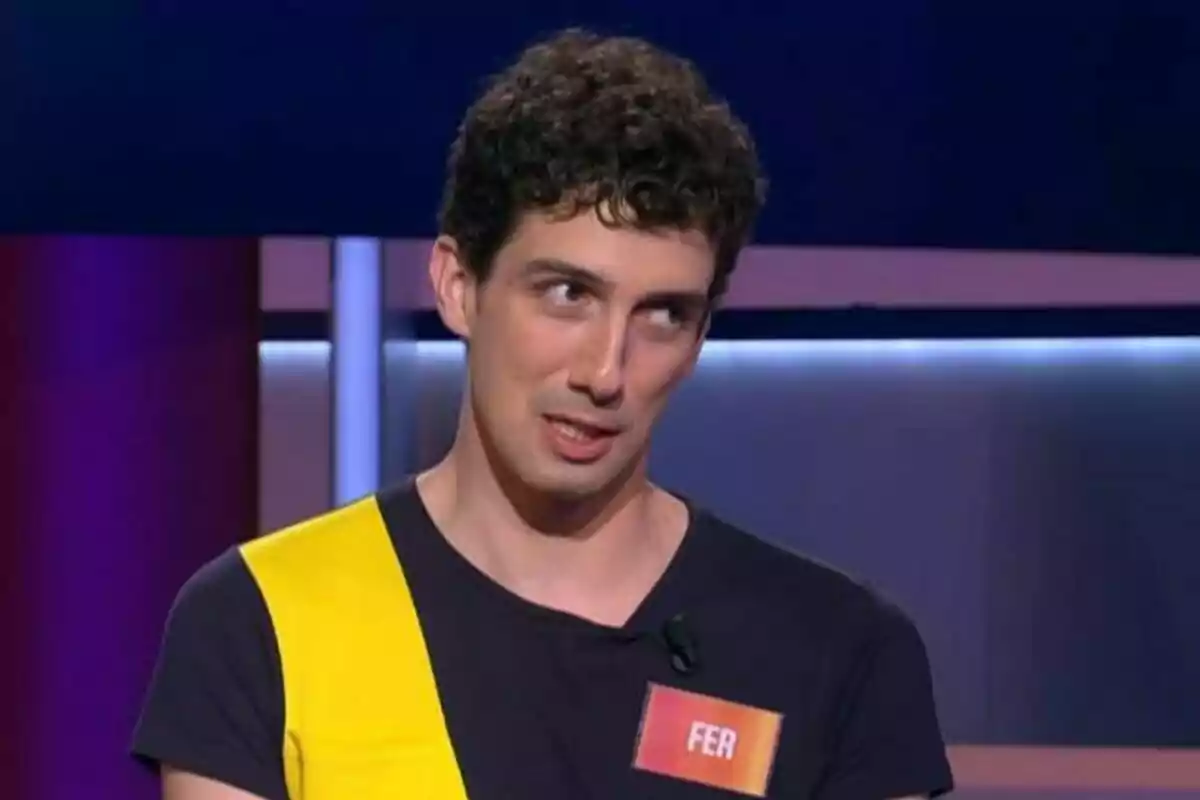 Fer Castro con cabello rizado y camiseta negra con una franja amarilla está en un set de Saber y Ganar y lleva una etiqueta con el nombre Fer. Fer Castro con cabello rizado y camiseta negra con una franja amarilla está en un set de Saber y Ganar y lleva una etiqueta con el nombre Fer.