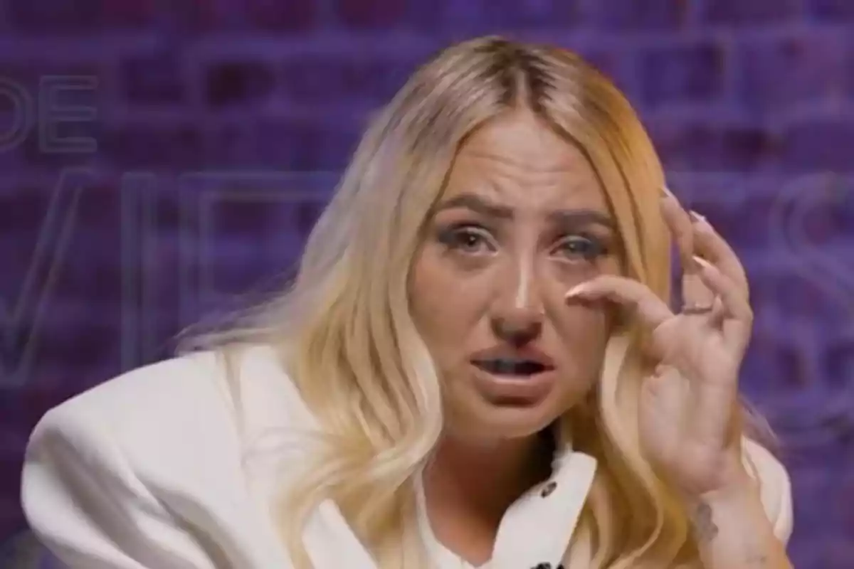 Rocío Flores rubia con expresión de tristeza y lágrimas en los ojos, vestida con saco blanco y fondo morado desenfocado