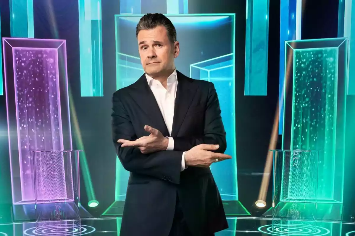 Iñaki López de traje oscuro y camisa blanca posando con los brazos cruzados y señalando hacia los lados en un set iluminado con luces y paneles de colores neón en Tesoro o cacharro Iñaki López de traje oscuro y camisa blanca posando con los brazos cruzados y señalando hacia los lados en un set iluminado con luces y paneles de colores neón en Tesoro o cacharro