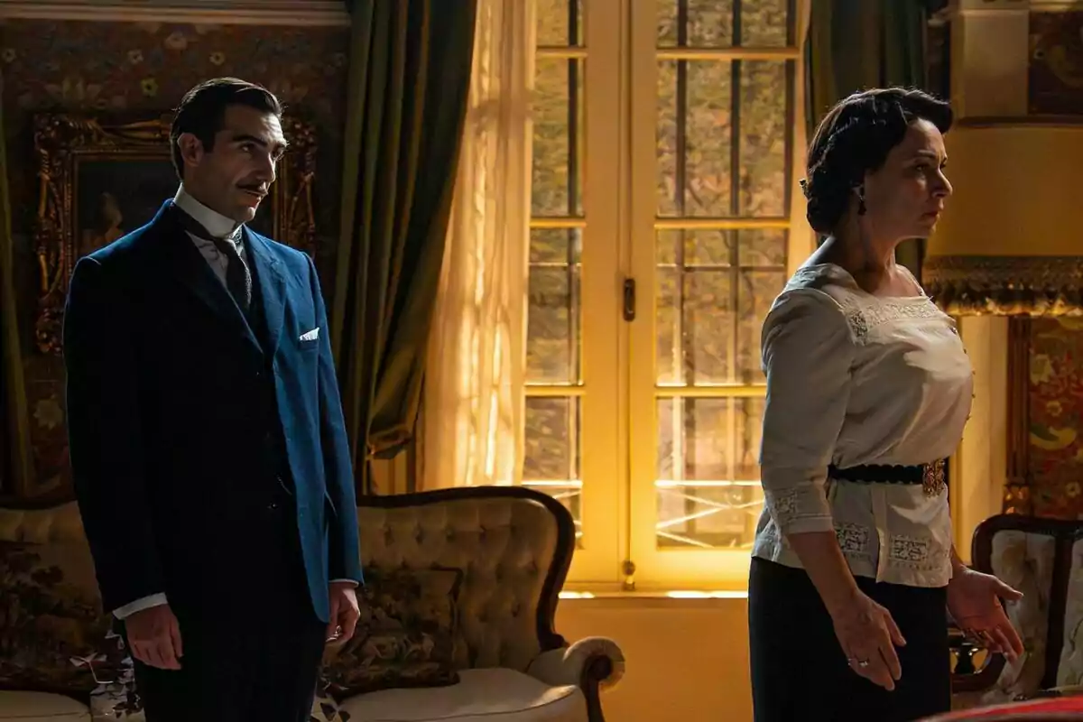 Guillermo Serrano amb bigoti i vestit blau fosc observa Isabel Serrano de cabell fosc i brusa clara que està dreta de perfil en una sala elegant amb cortines verdes i llum càlida entrant per una finestra gran a La Promesa. Guillermo Serrano amb bigoti i vestit blau fosc observa Isabel Serrano de cabell fosc i brusa clara que està dreta de perfil en una sala elegant amb cortines verdes i llum càlida entrant per una finestra gran a La Promesa.