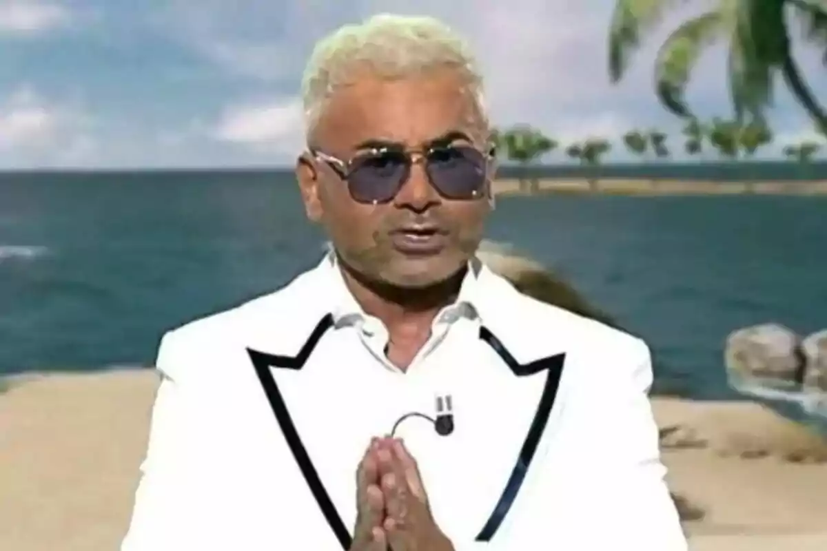 Jorge Javier Vázqeuez de cabello rubio y gafas de sol vistiendo un traje blanco con detalles negros, de pie frente a un fondo de playa con palmeras, juntando las manos en señal de saludo o agradecimiento en Supervivientes All Stars.