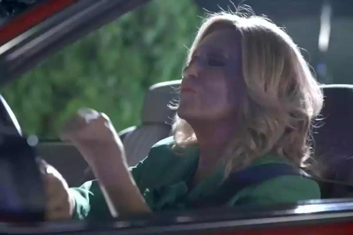 Lydia Lozano rubia sentada en un auto rojo haciendo un gesto de celebración con el puño cerrado en la promo de De Viernes