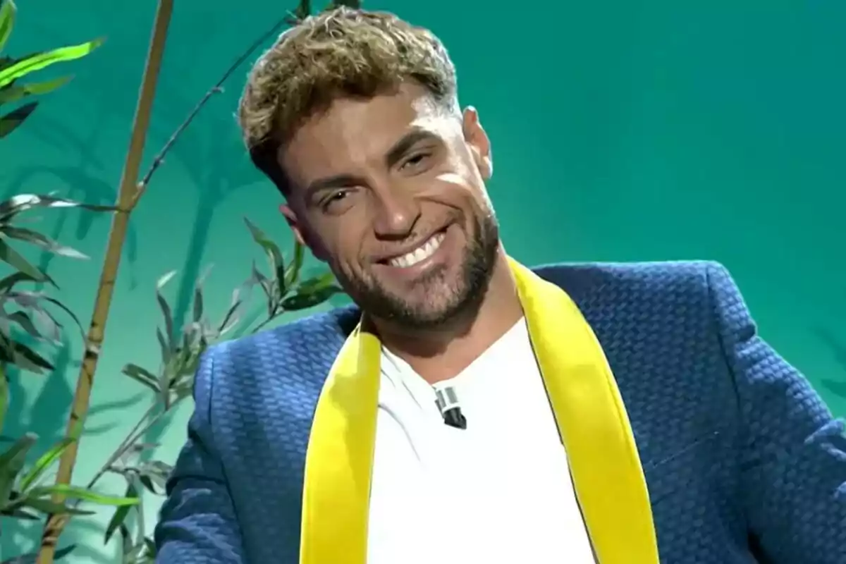 Montoya sonriente con chaqueta azul y bufanda amarilla sentado frente a un fondo verde con plantas decorativas en Supervivientes All Stars Montoya sonriente con chaqueta azul y bufanda amarilla sentado frente a un fondo verde con plantas decorativas en Supervivientes All Stars