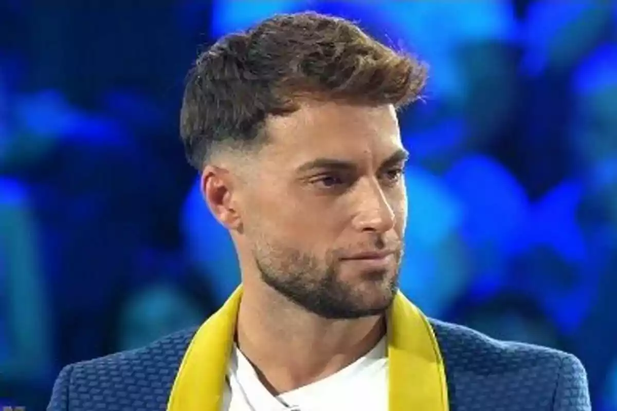 Montoya joven con barba y cabello corto, vestido con chaqueta azul y solapa amarilla, en un ambiente iluminado de color azul en Supervivientes