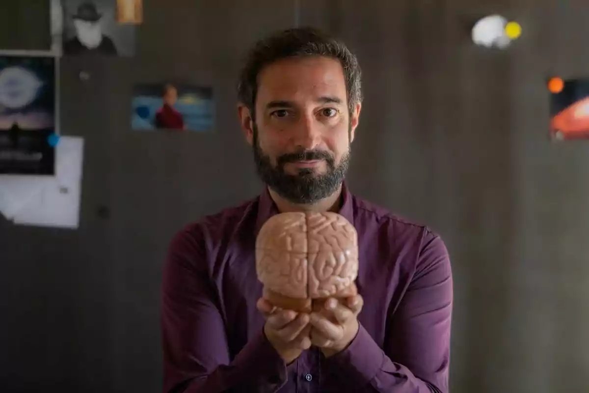 Pere Estupinyà de barba y camisa morada sostiene un modelo de cerebro de plástico frente a él con fondo desenfocado de pared y fotografías en El Cazador de Cerebros. Pere Estupinyà de barba y camisa morada sostiene un modelo de cerebro de plástico frente a él con fondo desenfocado de pared y fotografías en El Cazador de Cerebros.