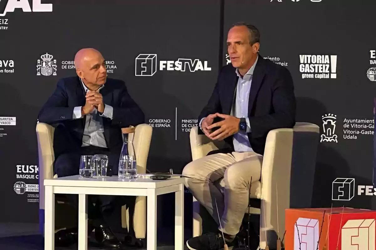 Jaime Guerra i Santi Acosta amb vestit conversen asseguts en butaques blanques sobre un escenari en la presentació d’El preu de... amb un fons negre ple de logotips institucionals i d’un festival; entre ells hi ha una taula amb gots i una gerra d’aigua.