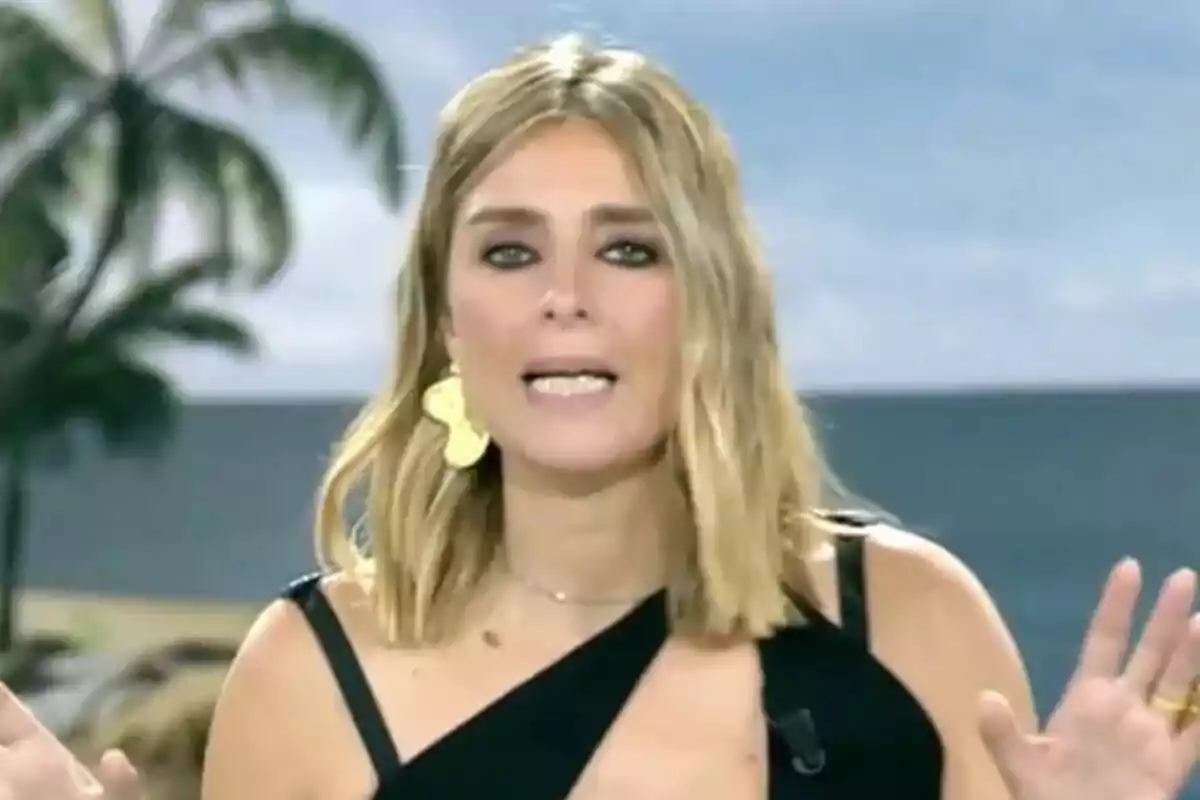 Sandra Barneda rossa amb vestit negre i arracades grans parlant davant d’una càmera amb fons de palmera i cel blau a Supervivientes All Stars