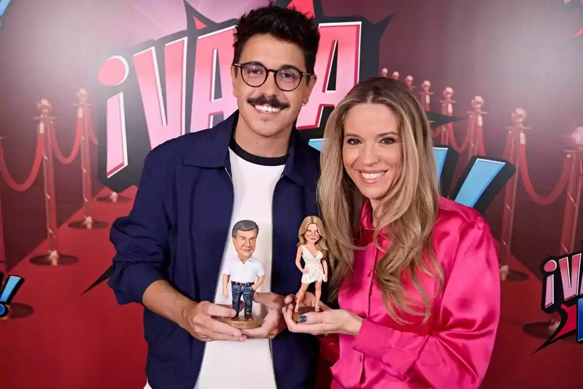 Cristina Lasvignes i Fran Ramírez somriuen davant d’un fons de catifa vermella i sostenen figures personalitzades que els representen a ¡Vaya Fama!.