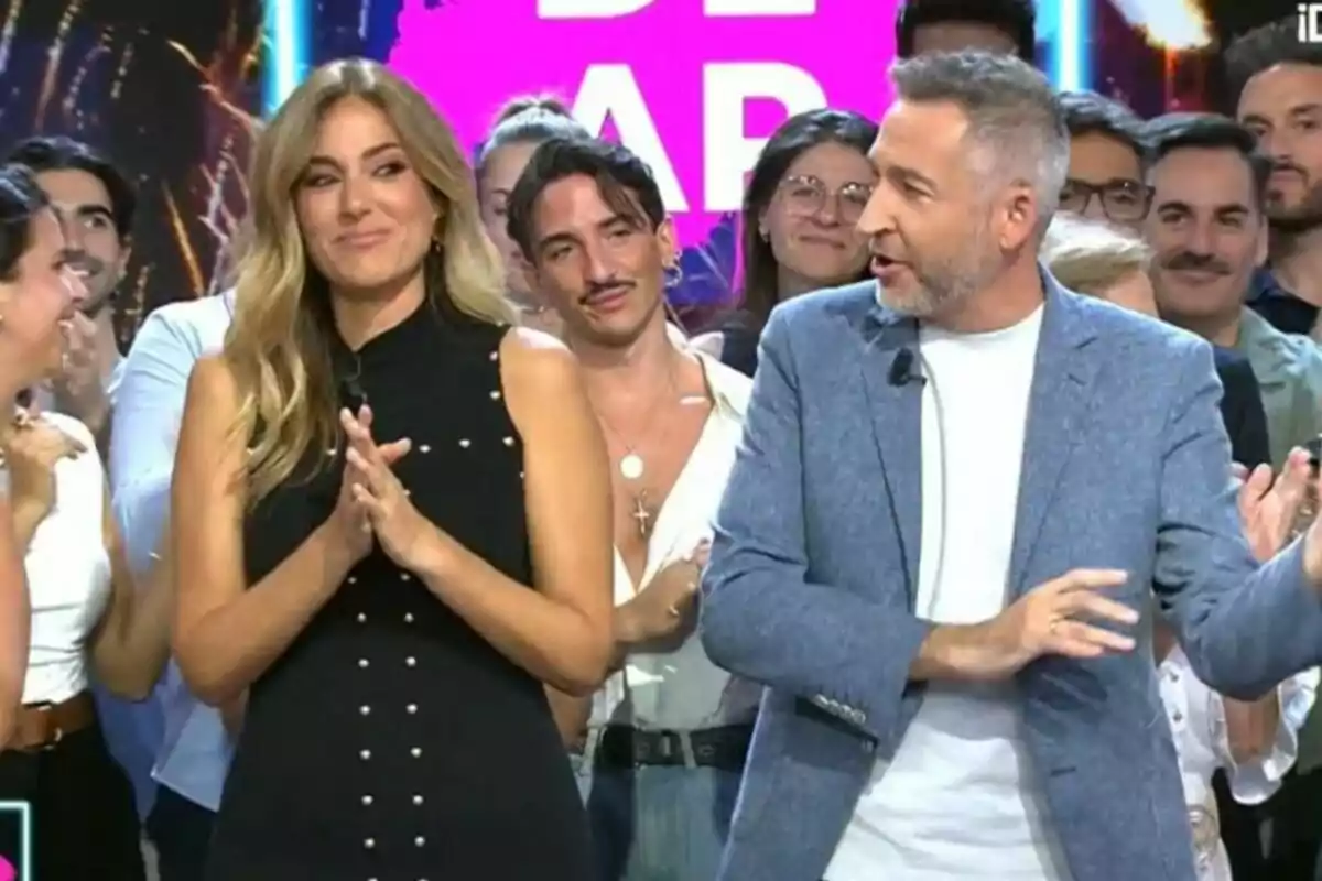 Un grupo de personas en un set de Tardear con Verónica Dulanto y Frank Blanco en primer plano, uno de ellos con vestido negro y el otro con chaqueta azul, rodeados de público sonriente y luces de fondo. Un grupo de personas en un set de Tardear con Verónica Dulanto y Frank Blanco en primer plano, uno de ellos con vestido negro y el otro con chaqueta azul, rodeados de público sonriente y luces de fondo.