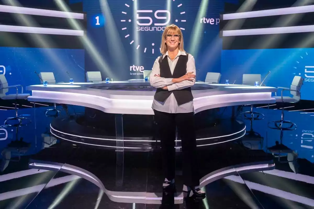 Gemma Nierga amb els braços plegats davant d'una taula rodona en un set de televisió amb el logo de Gemma Nierga amb els braços plegats davant d'una taula rodona en un set de televisió amb el logo de