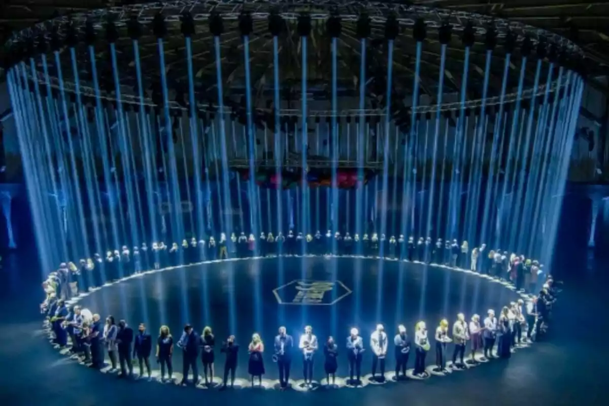 Un grupo de personas está de pie en un círculo iluminado por haces de luz azul que descienden desde el techo en un ambiente oscuro y futurista en 99 to Beat. Un grupo de personas está de pie en un círculo iluminado por haces de luz azul que descienden desde el techo en un ambiente oscuro y futurista en 99 to Beat.