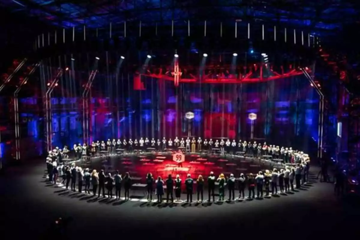 Un grup de persones està dret en un cercle sota llums de colors en un escenari ampli i fosc a 99 to Beat. Un grup de persones està dret en un cercle sota llums de colors en un escenari ampli i fosc a 99 to Beat.