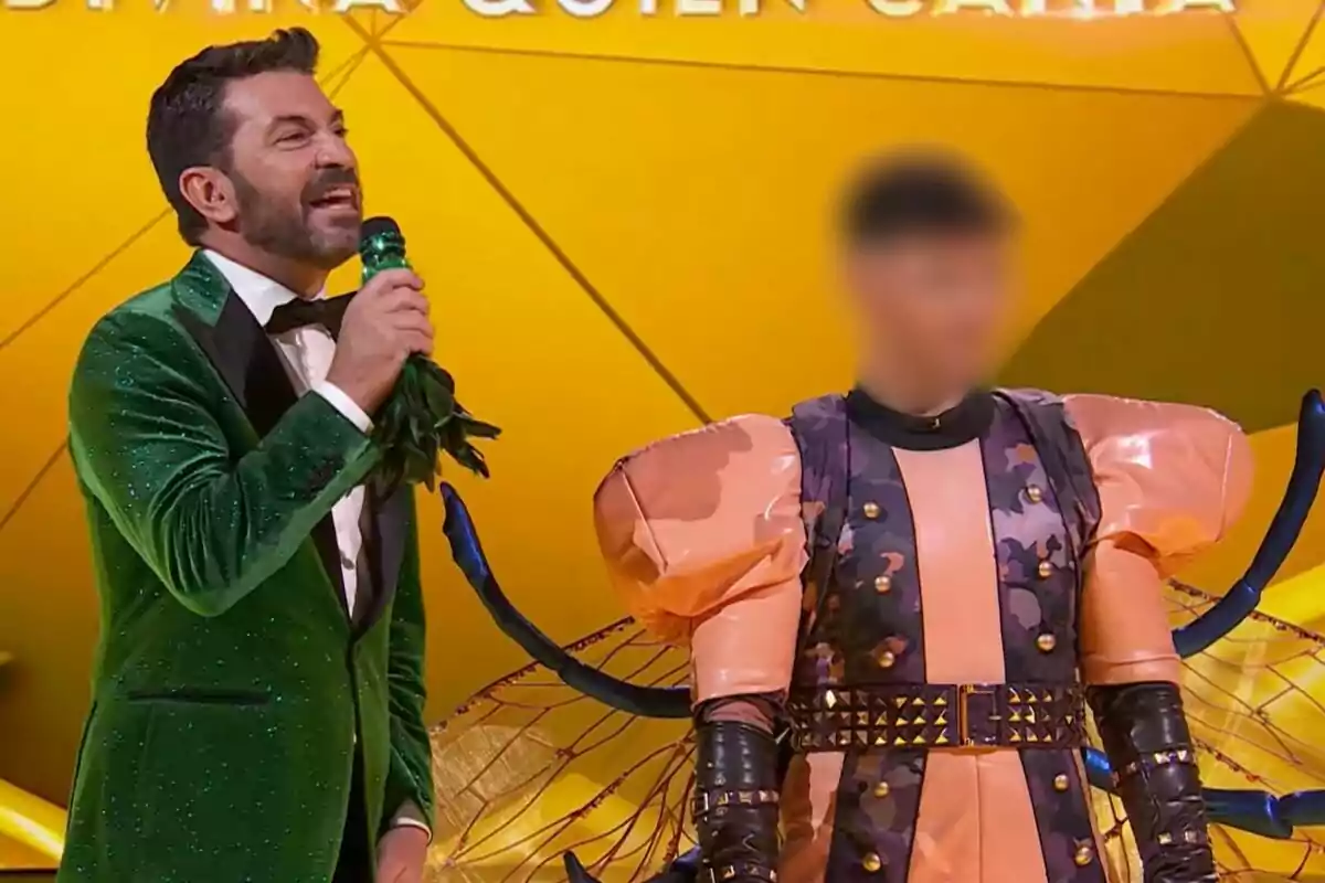 Arturo Valls con un traje verde brillante sostiene un micrófono mientras está al lado de una persona disfrazada con un traje colorido y voluminoso de Mosca en un escenario iluminado en Mask Singer.