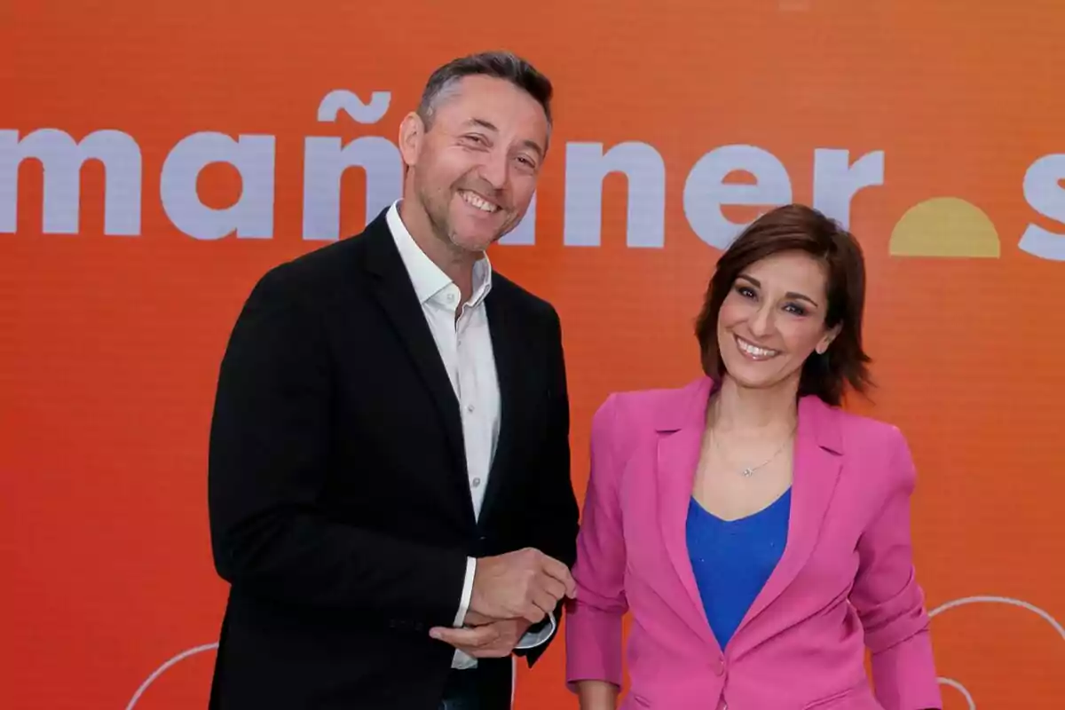 Javier Ruiz y Adela González sonríen frente a un fondo naranja con texto parcialmente visible que dice Mañaneros.