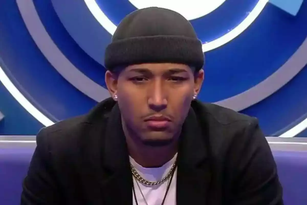 Adrián con gorro negro y chaqueta oscura está sentado frente a un fondo azul con círculos en Gran Hermano. Adrián con gorro negro y chaqueta oscura está sentado frente a un fondo azul con círculos en Gran Hermano.