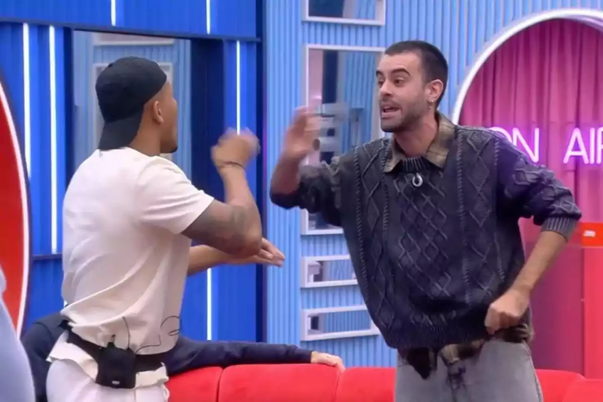 Adrián i Ruvens discutint a la casa de Gran Hermano amb llums de neó i un sofà vermell. Adrián i Ruvens discutint a la casa de Gran Hermano amb llums de neó i un sofà vermell.