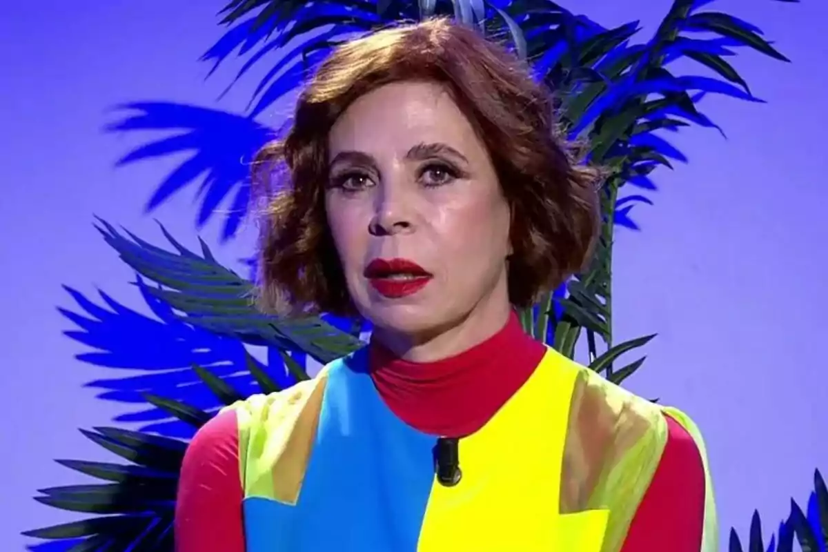 Ágatha Ruiz de la Prada con cabello corto y castaño, vestida con un atuendo colorido, está frente a un fondo azul con hojas de plantas en De Viernes.