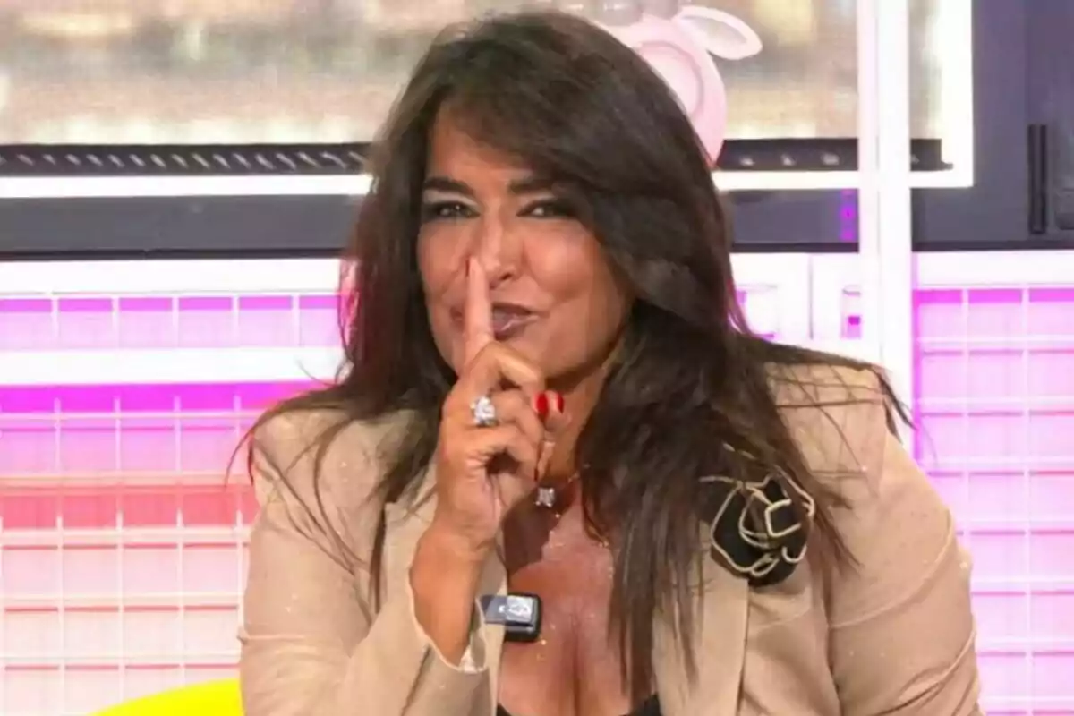 Aída Nizar con cabello oscuro haciendo gesto de silencio con el dedo índice frente a la boca, vestida con chaqueta beige y fondo colorido en Ni que fuéramos. Aída Nizar con cabello oscuro haciendo gesto de silencio con el dedo índice frente a la boca, vestida con chaqueta beige y fondo colorido en Ni que fuéramos.
