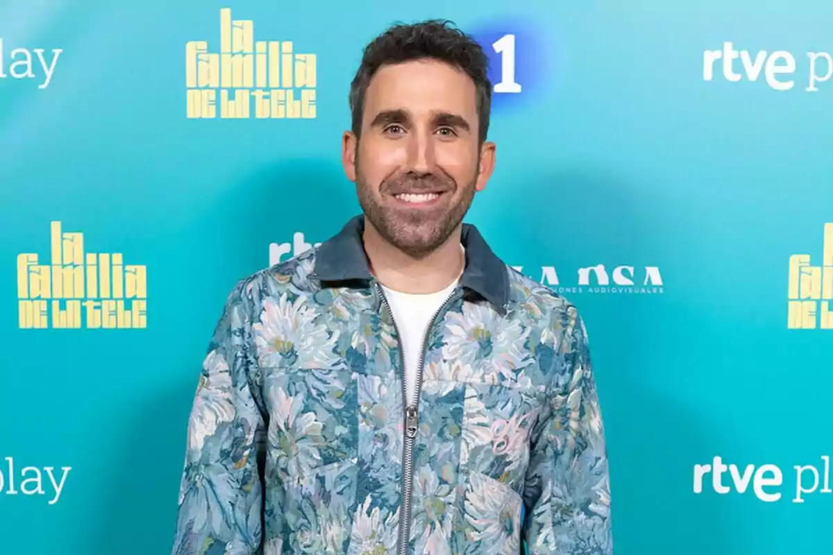 Aitor Albizua somrient amb barba i cabell fosc porta una jaqueta amb estampat floral sobre una samarreta blanca, posant davant d'un fons blau amb logotips de 