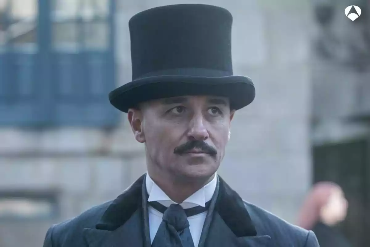 Alain Hernández con sombrero de copa y bigote, vestido con traje formal, mira hacia adelante con expresión seria en Las hijas de la criada. Alain Hernández con sombrero de copa y bigote, vestido con traje formal, mira hacia adelante con expresión seria en Las hijas de la criada.