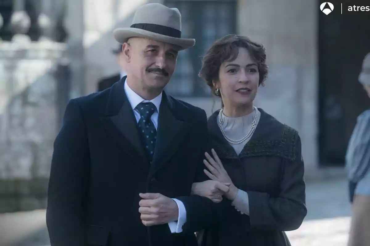Alain Hernández y Verónica Sánchez vestidos con ropa de época, el hombre lleva sombrero y traje oscuro, mientras que la mujer lleva un vestido elegante y collares de perlas, ambos están de pie juntos al aire libre en Las hijas de la criada. Alain Hernández y Verónica Sánchez vestidos con ropa de época, el hombre lleva sombrero y traje oscuro, mientras que la mujer lleva un vestido elegante y collares de perlas, ambos están de pie juntos al aire libre en Las hijas de la criada.